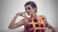 Mulher de cabelo curto e undercut, blusa xadrez laranja, pose reflexiva, mão no rosto. Fundo claro e minimalista.