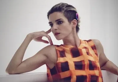 Mulher de cabelo curto e undercut, blusa xadrez laranja, pose reflexiva, mão no rosto. Fundo claro e minimalista.