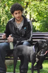 Jovem de barba e cabelo encaracolado sentado num banco de parque com um cão preto de coleira. Ele segura uma garrafa.