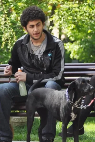 Jovem de barba e cabelo encaracolado sentado num banco de parque com um cão preto de coleira. Ele segura uma garrafa.