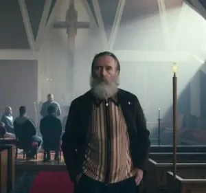 Homem barbudo sério em destaque numa igreja com cruz, enquanto outros homens estão sentados ao fundo. Luz dramática.
