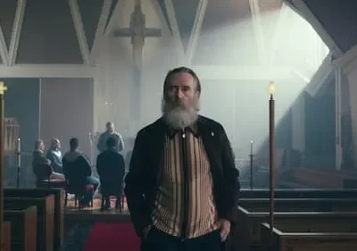 Homem barbudo sério em destaque numa igreja com cruz, enquanto outros homens estão sentados ao fundo. Luz dramática.