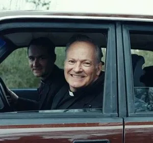 Quatro homens sorridentes, incluindo dois padres, dentro de um carro. O clima é alegre e amigável. Um terço pendura no espelho.