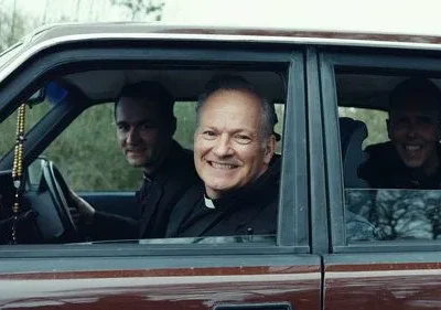 Quatro homens sorridentes, incluindo dois padres, dentro de um carro. O clima é alegre e amigável. Um terço pendura no espelho.