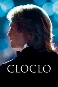 Homem de perfil com cabelo claro e volumoso,iluminado por trás,em terno preto,olhando para luzes azuis desfocadas.