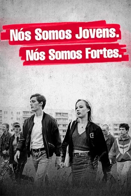 Jovens de semblante sério,um casal de mãos dadas,caminham em um campo. Cenário urbano cinza,pôster P&B com texto vermelho.