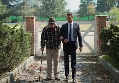 Jovem de terno apoia idoso com chapéu e bengala, caminhando em trilha de jardim com portão branco ao fundo. Clima de apoio.