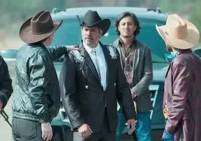 Grupo de homens de chapéu de cowboy, um com rifle, em frente a um SUV escuro. O central usa terno bordado e parece sério. Cenário rústico.