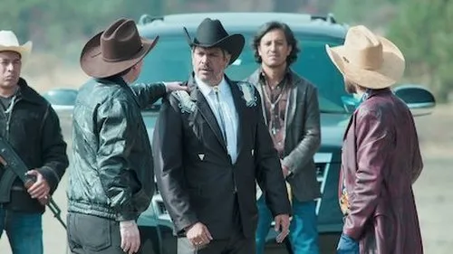 Grupo de homens de chapéu de cowboy,um com rifle,em frente a um SUV escuro. O central usa terno bordado e parece sério. Cenário rústico.