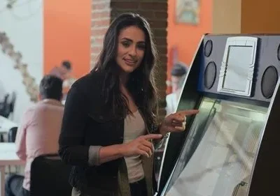 Mulher sorridente com jaqueta escura aponta para uma jukebox. Pessoas e mesas desfocadas no fundo de um restaurante.
