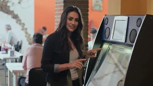 Mulher sorridente com jaqueta escura aponta para uma jukebox. Pessoas e mesas desfocadas no fundo de um restaurante.