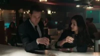 Um homem de terno e uma mulher de cabelo escuro sentados num bar,ambos com copos de shot. Ambiente escuro e pensativo.