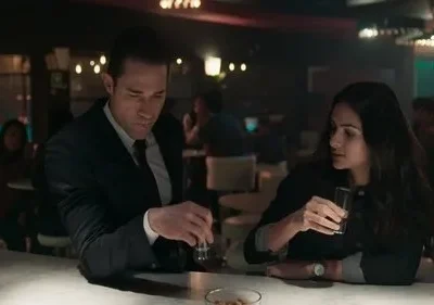 Um homem de terno e uma mulher de cabelo escuro sentados num bar, ambos com copos de shot. Ambiente escuro e pensativo.