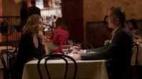 Mulher e homem em jantar íntimo num restaurante. Ela pensativa,em preto. Ele sorri de terno. Ambiente acolhedor,à meia-luz.