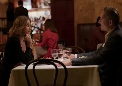 Mulher e homem em jantar íntimo num restaurante. Ela pensativa, em preto. Ele sorri de terno. Ambiente acolhedor, à meia-luz.