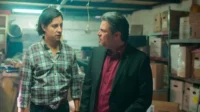 Dois homens sérios,um de xadrez e outro de terno,num armazém com caixas. Eles se olham com tensão.