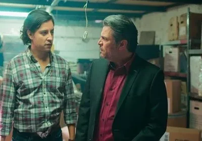 Dois homens sérios, um de xadrez e outro de terno, num armazém com caixas. Eles se olham com tensão.