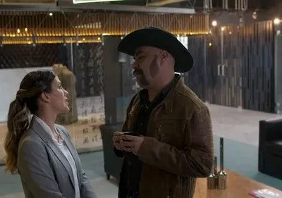 Uma mulher sorridente em blazer cinza conversa com um homem de chapéu cowboy. Estão num ambiente moderno com caligrafia asiática e iluminação acolhedora.