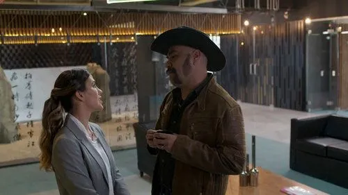 Uma mulher sorridente em blazer cinza conversa com um homem de chapéu cowboy. Estão num ambiente moderno com caligrafia asiática e iluminação acolhedora.