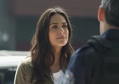 Mulher de cabelo escuro olha para homem de uniforme policial, visto de costas. Clima sério e urbano.
