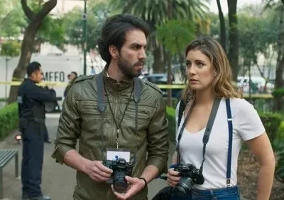 Dois jornalistas com câmeras observam uma área isolada por fita amarela num parque, com policiais e uma van. Clima tenso.