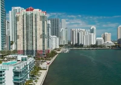 Paisagem urbana ensolarada: prédios altos e modernos à beira de um mar azul, com palmeiras e céu claro.