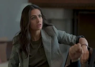 Uma mulher de cabelo escuro e blazer xadrez, com expressão atenta e séria, olha para o lado num interior. Clima dramático e intenso.
