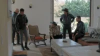 Cinco homens sérios em uma sala moderna com janelão para a natureza. Dois usam chapéu,três vestem jaquetas de couro escuras.