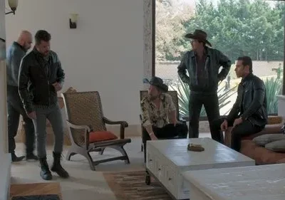 Cinco homens sérios em uma sala moderna com janelão para a natureza. Dois usam chapéu, três vestem jaquetas de couro escuras.