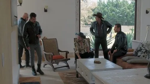 Cinco homens sérios em uma sala moderna com janelão para a natureza. Dois usam chapéu,três vestem jaquetas de couro escuras.