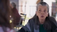 Mulher idosa de cabelos grisalhos,com olhar atento e boca aberta,usa um xale em um pátio ensolarado.