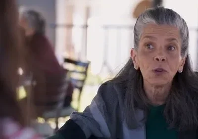 Mulher idosa de cabelos grisalhos, com olhar atento e boca aberta, usa um xale em um pátio ensolarado.