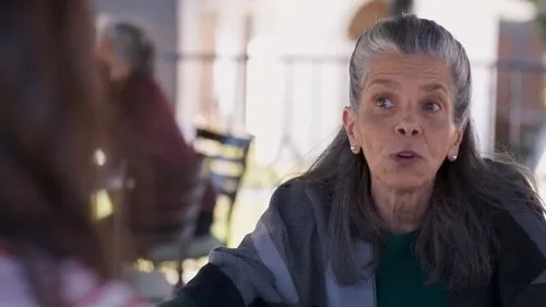 Mulher idosa de cabelos grisalhos,com olhar atento e boca aberta,usa um xale em um pátio ensolarado.