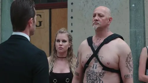 Homem de terno de costas encara careca tatuado e mulher loira de maquiagem escura. Ambiente sério,de confronto.