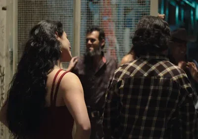 Mulher de cabelo escuro e vestido vermelho conversa com grupo em rua noturna com grafites. Cena urbana animada.
