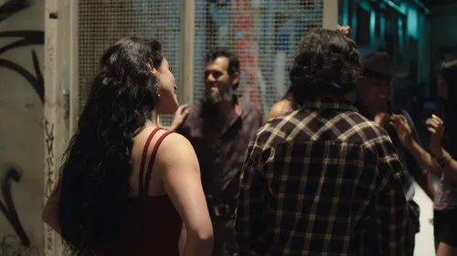 Mulher de cabelo escuro e vestido vermelho conversa com grupo em rua noturna com grafites. Cena urbana animada.