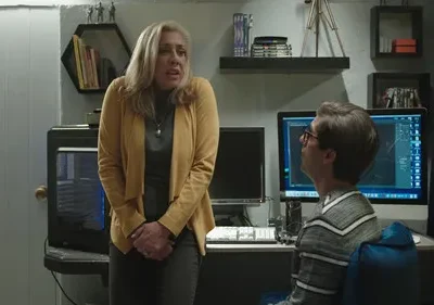 Mulher loira em amarelo, parecendo aflita, fala com jovem de óculos sentado ao computador em ambiente de tecnologia.