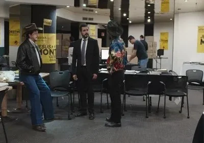 Três homens se destacam: um de chapéu e jaqueta, um de terno e um de camisa colorida, em sala com banners. Outras pessoas trabalham.