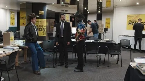 Três homens se destacam:um de chapéu e jaqueta,um de terno e um de camisa colorida,em sala com banners. Outras pessoas trabalham.