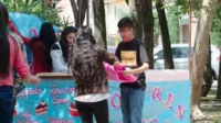 O Último Dragão – T01E31:Episódio 31 7 Jovens montam barraca de doces azul com cupcakes em evento ao ar livre. Rapaz de óculos segura tecido rosa;mulheres no balcão.