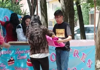 Jovens montam barraca de doces azul com cupcakes em evento ao ar livre. Rapaz de óculos segura tecido rosa; mulheres no balcão.