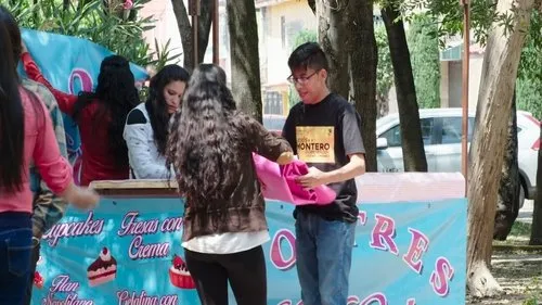 Jovens montam barraca de doces azul com cupcakes em evento ao ar livre. Rapaz de óculos segura tecido rosa; mulheres no balcão.