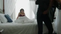 O Último Dragão – T01E33:Episódio 33 5 Mulher em sutiã na cama,com expressão surpresa,olha para pessoa se vestindo. Quarto claro,clima de intimidade e tensão.