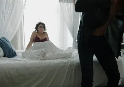 Mulher em sutiã na cama, com expressão surpresa, olha para pessoa se vestindo. Quarto claro, clima de intimidade e tensão.
