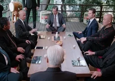 Grupo de homens sérios em ternos sentados em torno de uma mesa baixa num ambiente japonês. Reunião formal e tensa.