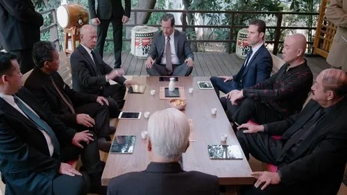 Grupo de homens sérios em ternos sentados em torno de uma mesa baixa num ambiente japonês. Reunião formal e tensa.