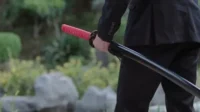 O Último Dragão – T01E36:Episódio 36 2 Homem de terno preto segura katana com cabo vermelho e bainha preta,em cenário verde de jardim. Tom sério e misterioso.