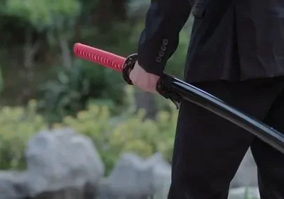Homem de terno preto segura katana com cabo vermelho e bainha preta, em cenário verde de jardim. Tom sério e misterioso.