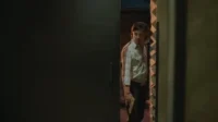 O Último Dragão – T01E37:Episódio 37 1 Homem de camisa clara e calça escura,segurando uma pistola dourada,espreita numa abertura escura com olhar tenso.