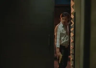 Homem de camisa clara e calça escura, segurando uma pistola dourada, espreita numa abertura escura com olhar tenso.